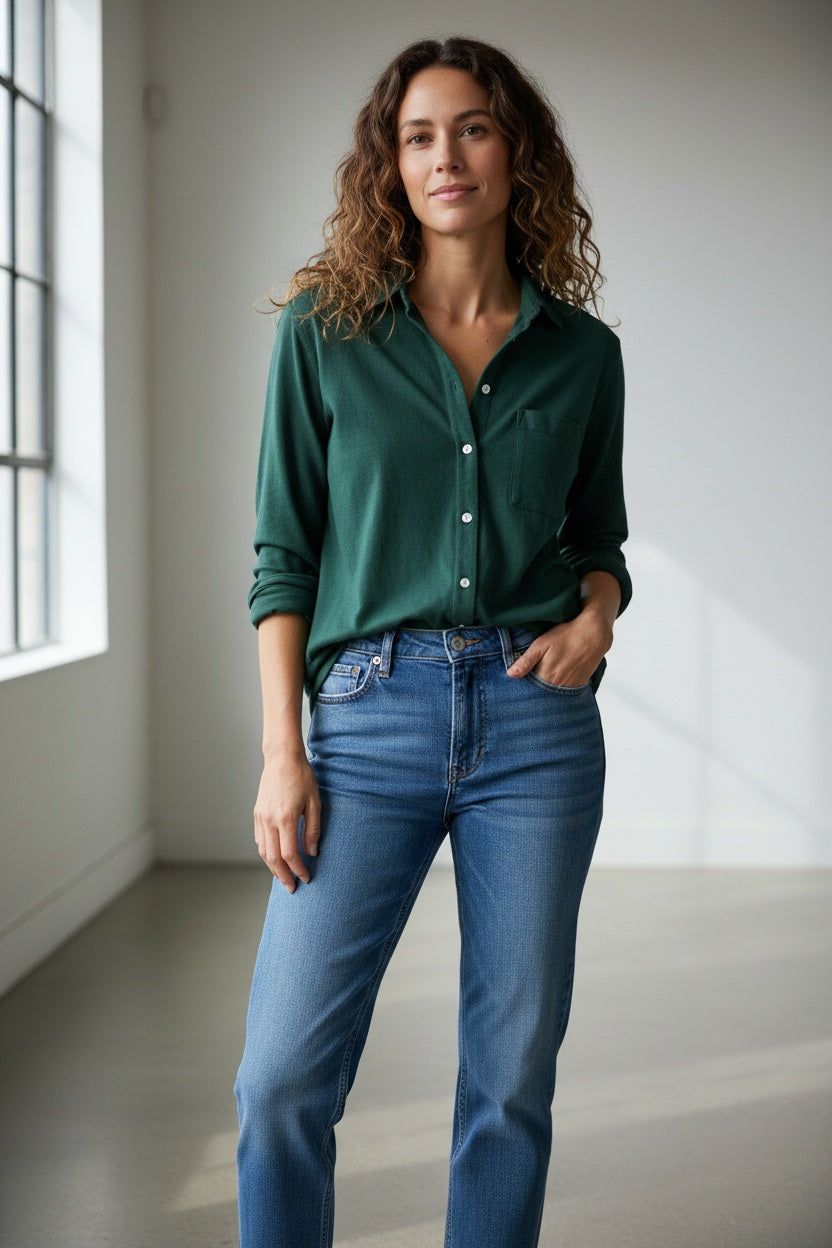 A woman with curly hair, smiling softly by a window indoors with one hand in her pocket, wears blue jeans and the FRANK & EILEEN Waverley Button Up Tee in Vintage Jersey Pine.