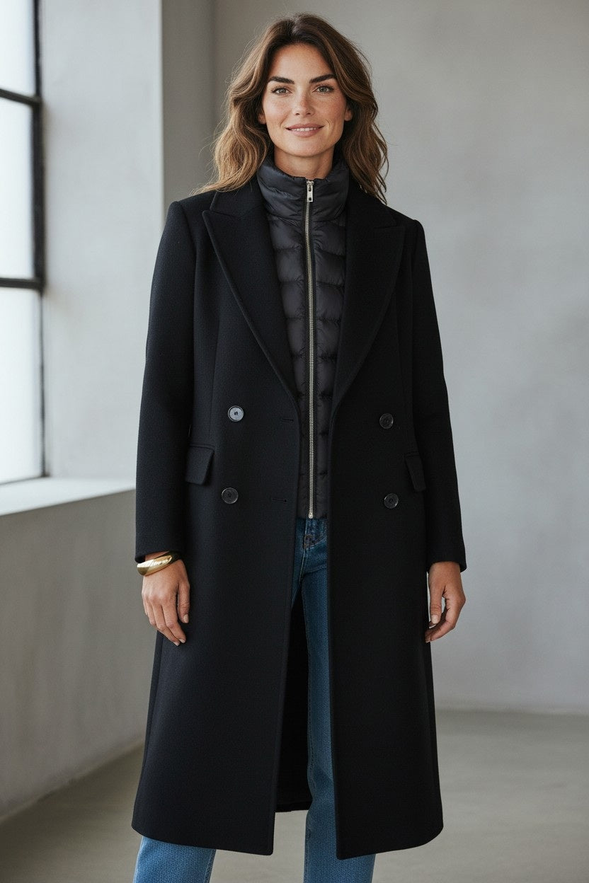 A woman stands indoors by a large window, smiling at the camera, wearing blue jeans and the KINROSS CASHMERE Quilted Insert Notch Collar Coat in black layered over a quilted black jacket.