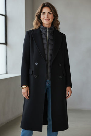 A woman stands indoors by a large window, smiling at the camera, wearing blue jeans and the KINROSS CASHMERE Quilted Insert Notch Collar Coat in black layered over a quilted black jacket.