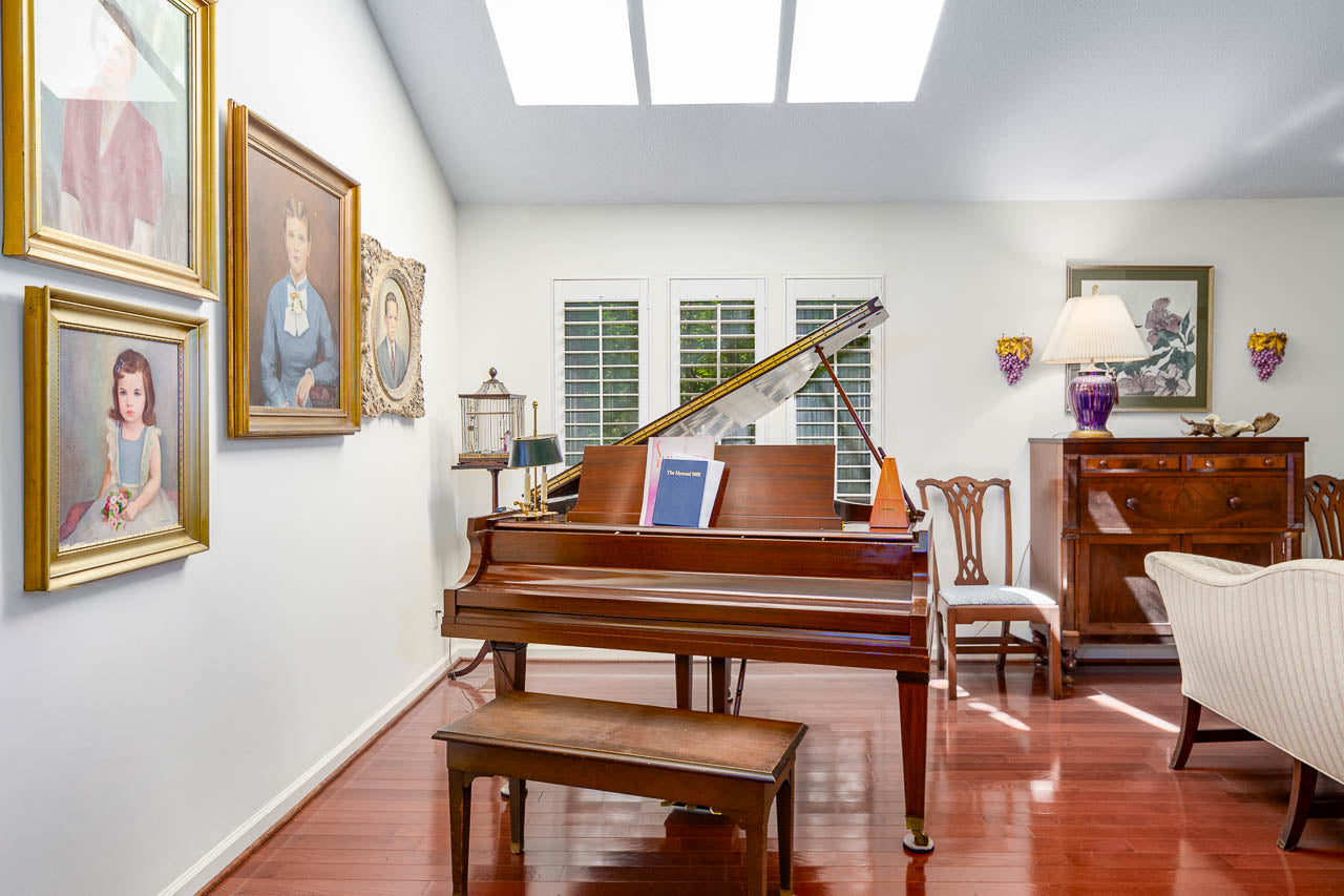 A grand piano with a bench sits in a bright room with hardwood floors, white walls, framed portraits, a wooden cabinet, chairs, a lamp, and large skylights overhead letting in natural light.