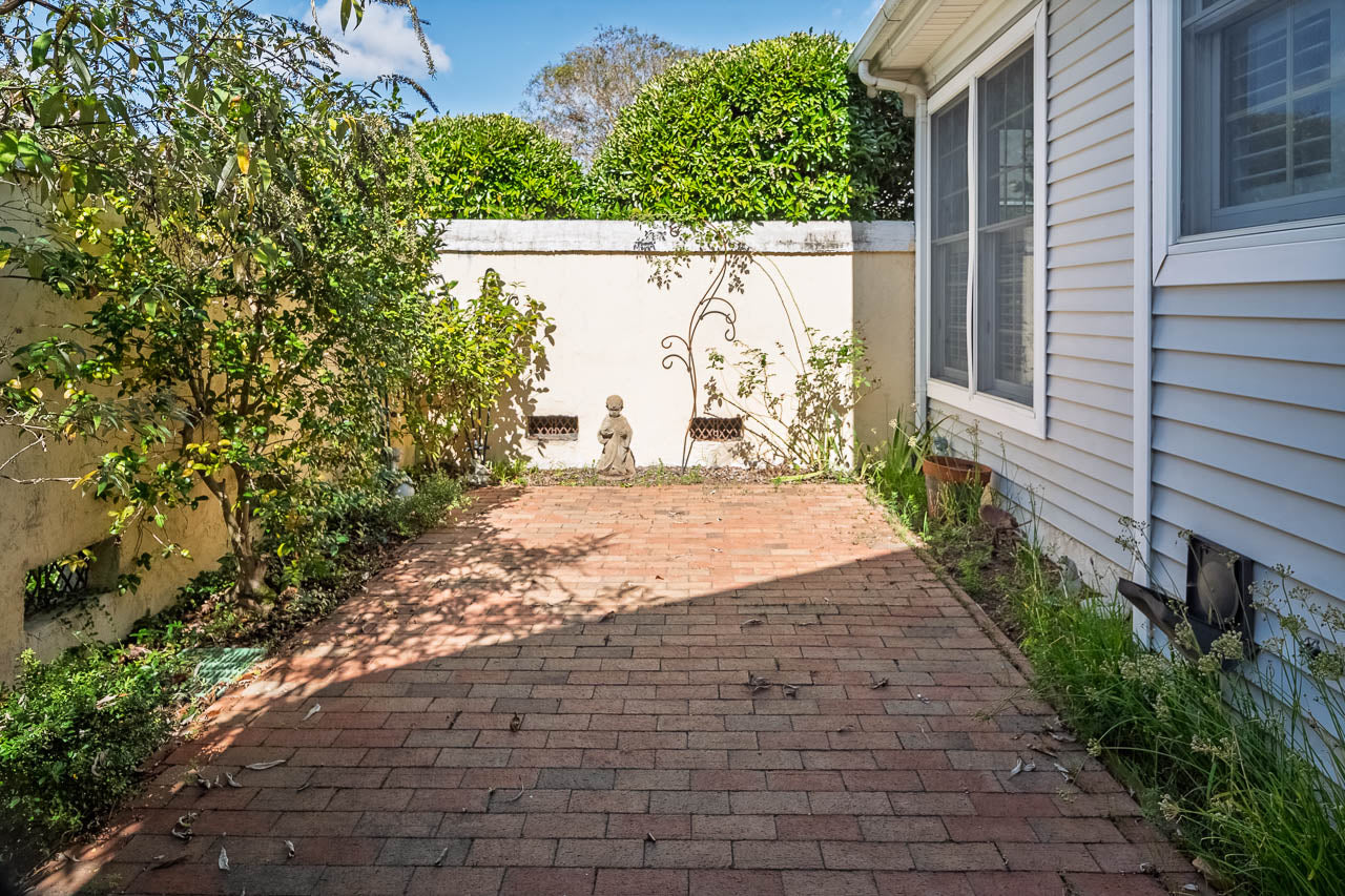 A small brick patio bordered by plants, with a white wall at the end featuring a statue and wall art. A window is visible on the right side of a light-colored house.