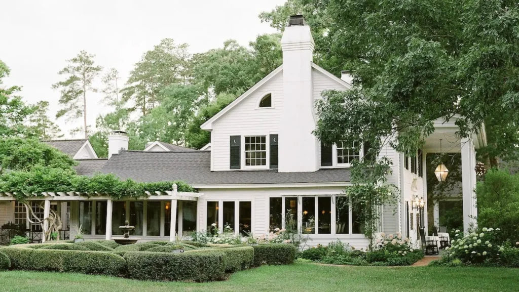 A large white house with black shutters, a tall chimney, and a covered porch surrounded by lush green trees, manicured hedges, a trellis, and a central fountain in the garden.