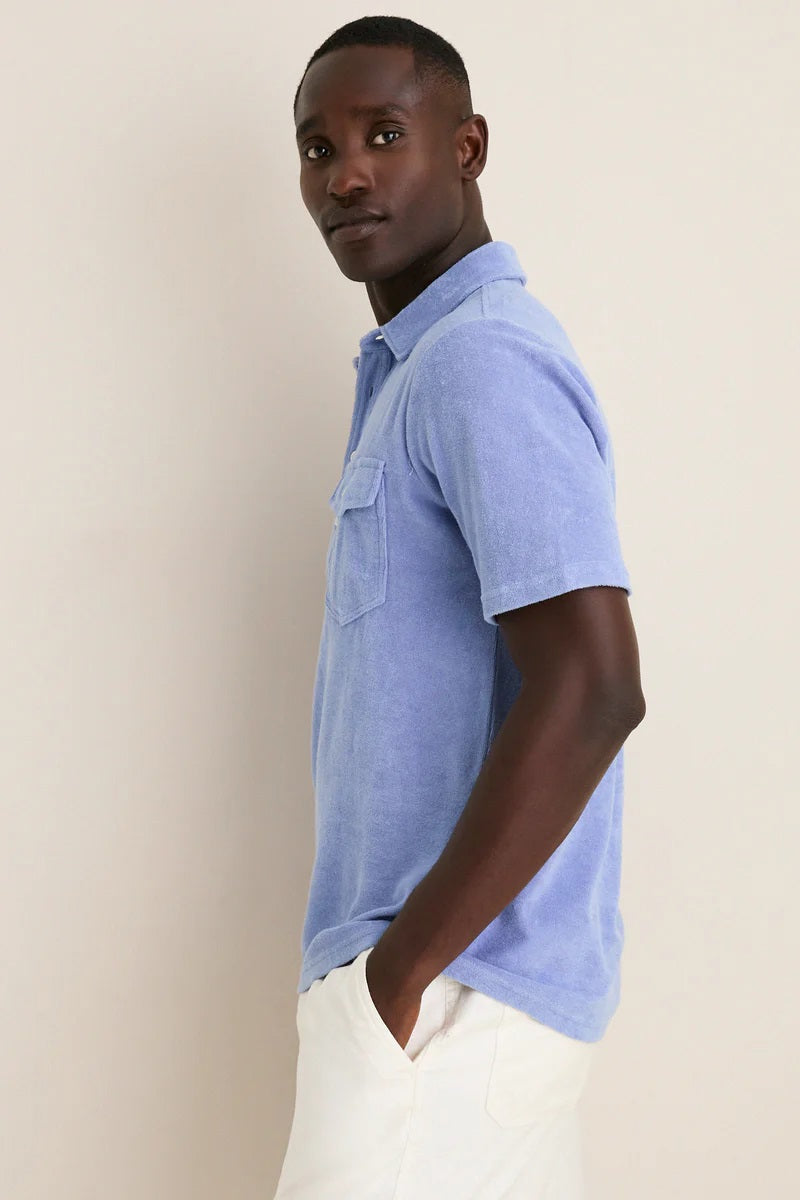 A person in a periwinkle Criquet Terrycloth Players Shirt and white shorts stands facing away from the camera against a plain beige background.