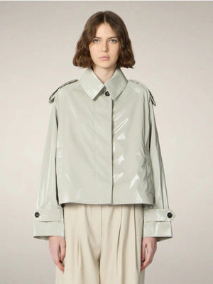 A person with chin-length brown hair wears the SAVE THE DUCK - GAIA JACKET by SAVE THE DUCK, a shiny light-gray, cropped trench-style women's jacket over a white top, paired with high-waisted beige trousers and beige shoes against a plain background.