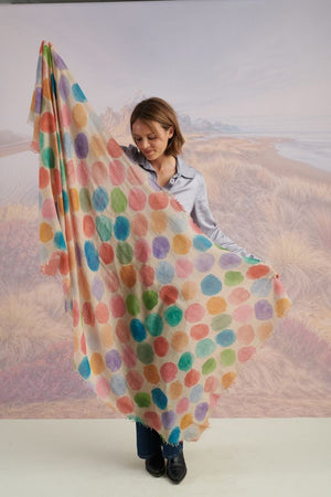 A woman with blonde hair displays the STORIATIPIC - BETTY WOOL SCARF, a large rectangular wool accessory with earth-toned polka dots, set against a scenic mountain landscape.