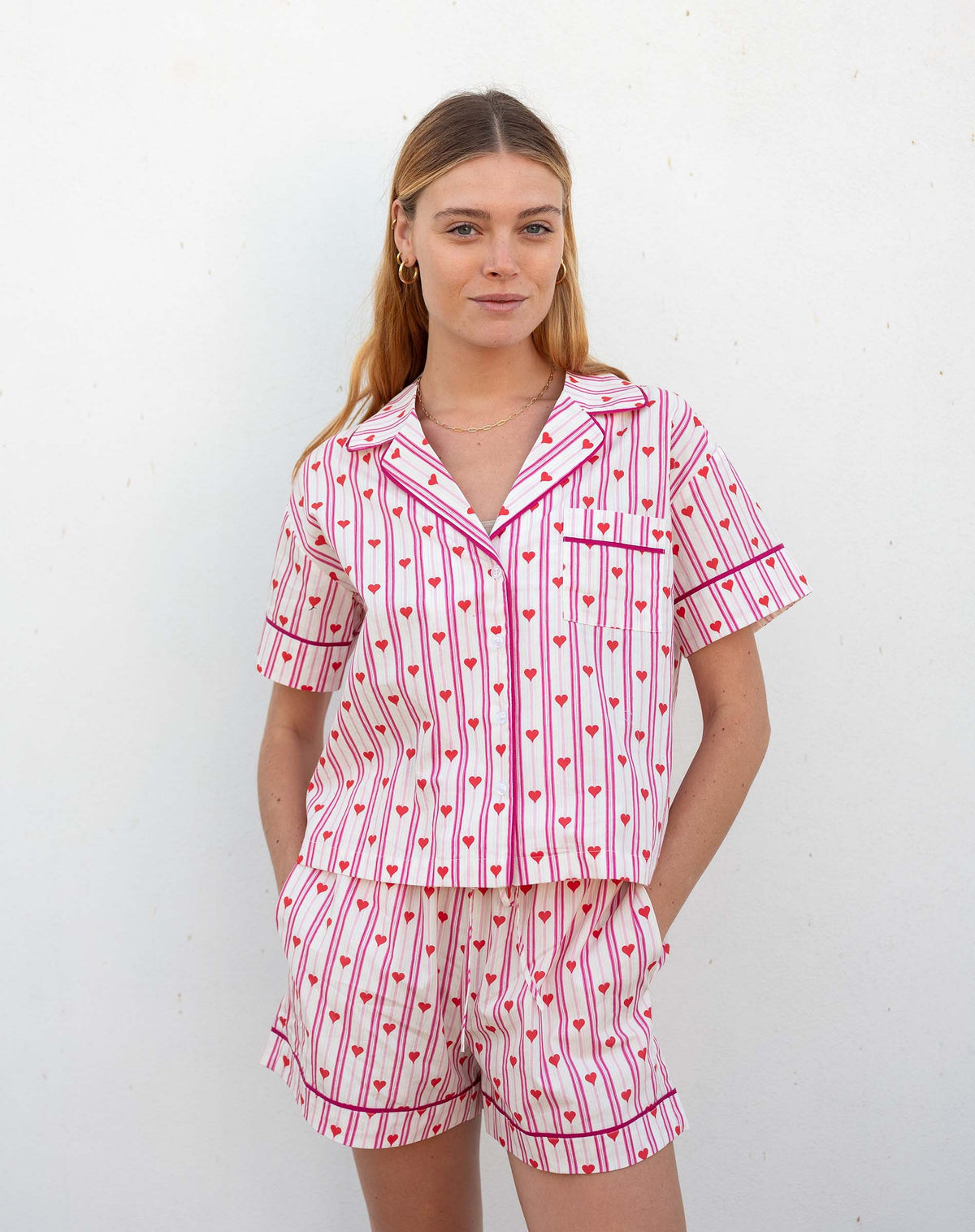 A woman with light brown hair smiles in MERSEA&#39;s OVER THE MOON PAJAMA SHORT SET LOVE NOTES— a heart print cotton short-sleeve button-up top and shorts—standing against a white background.