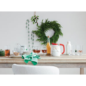 A GLASS BOWL WITH EMBEDDED HOLIDAY ICON filled with granola sits on a wooden table next to a Santa-shaped pitcher and artisan glass tumblers adorned with multicolor icon embellishments.