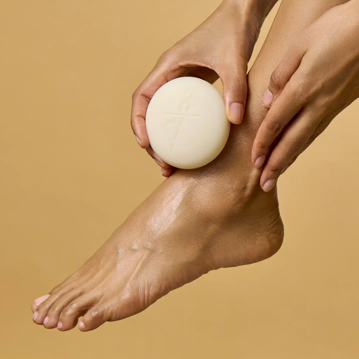 A person uses KATE MCLEOD’s THE FOOT STONE SOLID FOOT MOISTURIZER on their foot, holding the off-white round bar with one hand while the other rests on their ankle. The solid beige background emphasizes care for dry, cracked heels.