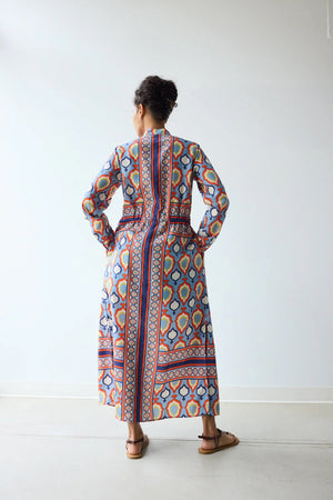 In a minimal room, a person with short curly hair and brown sandals exudes confidence in the HOPE FOR FLOWERS BAND COLLAR SHIRTDRESS IN CHAMBRAY MEDALLION, showcasing modern style from the HOPE FOR FLOWERS brand.