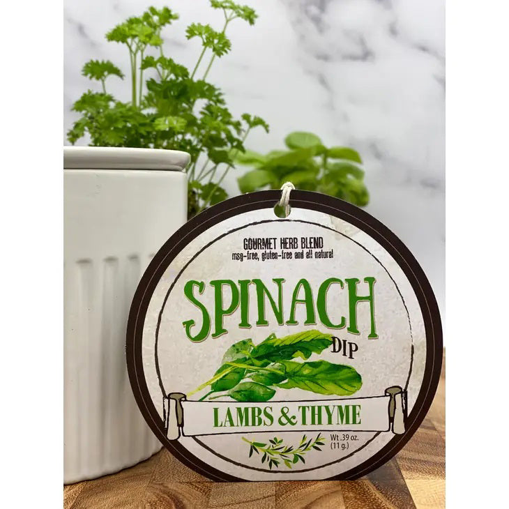 A circular label displays &quot;LAMBS &amp; THYME - SPINACH DIP&quot; alongside an illustration of spinach leaves. This easily prepared greeny goodness is contained in a white package, surrounded by fresh green herbs, likely parsley, set against a blurred background.