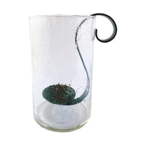 Three upright books are beside the CREATIVE COOP RECYCLED BUBBLE GLASS HURRICANE WITH METAL VOTIVE HOLDER, which contains a lit white candle and small pebbles. The lantern features a black metal handle and sits against a plain light background.