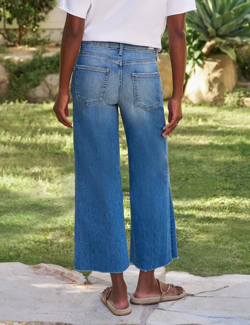 A person stands outdoors on grass, wearing the FRANK AND EILEEN - GALWAY WIDE LEG JEAN IN 2002 MID WASH and a white shirt, with their right hand relaxed by their side.