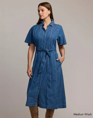 A woman wears the HINSON WU - JOANNA SHORT SLEEVE DENIM DRESS in medium wash, featuring a lightweight midi cut, short sleeves, front buttons, matching waist tie, and pockets. She has long brown hair and looks to the side. "Medium Wash" appears in the corner.