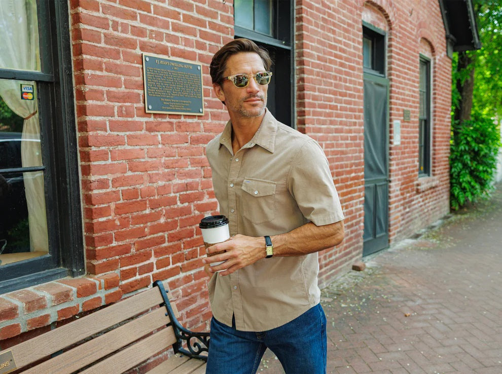 A man wearing the CRIQUET Short Sleeve Corduroy Pearl Snap Shirt in Irish Coffee stands in front of a dark door and red brick wall, holding a black object at chest level.