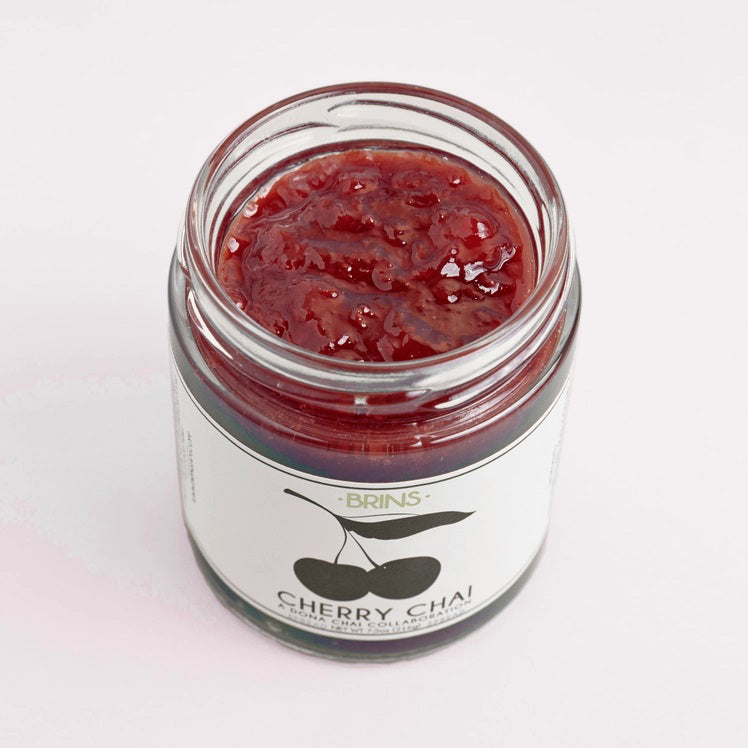 A glass jar of BRINS - CHERRY CHAI SPREAD & PRESERVE features a black lid and a label adorned with an illustration of two cherries alongside flavor details and collaboration notes. Made by BRINS with locally sourced New York sour cherries, this preserve is expertly infused with aromatic chai spices for a distinctive and flavorful experience.