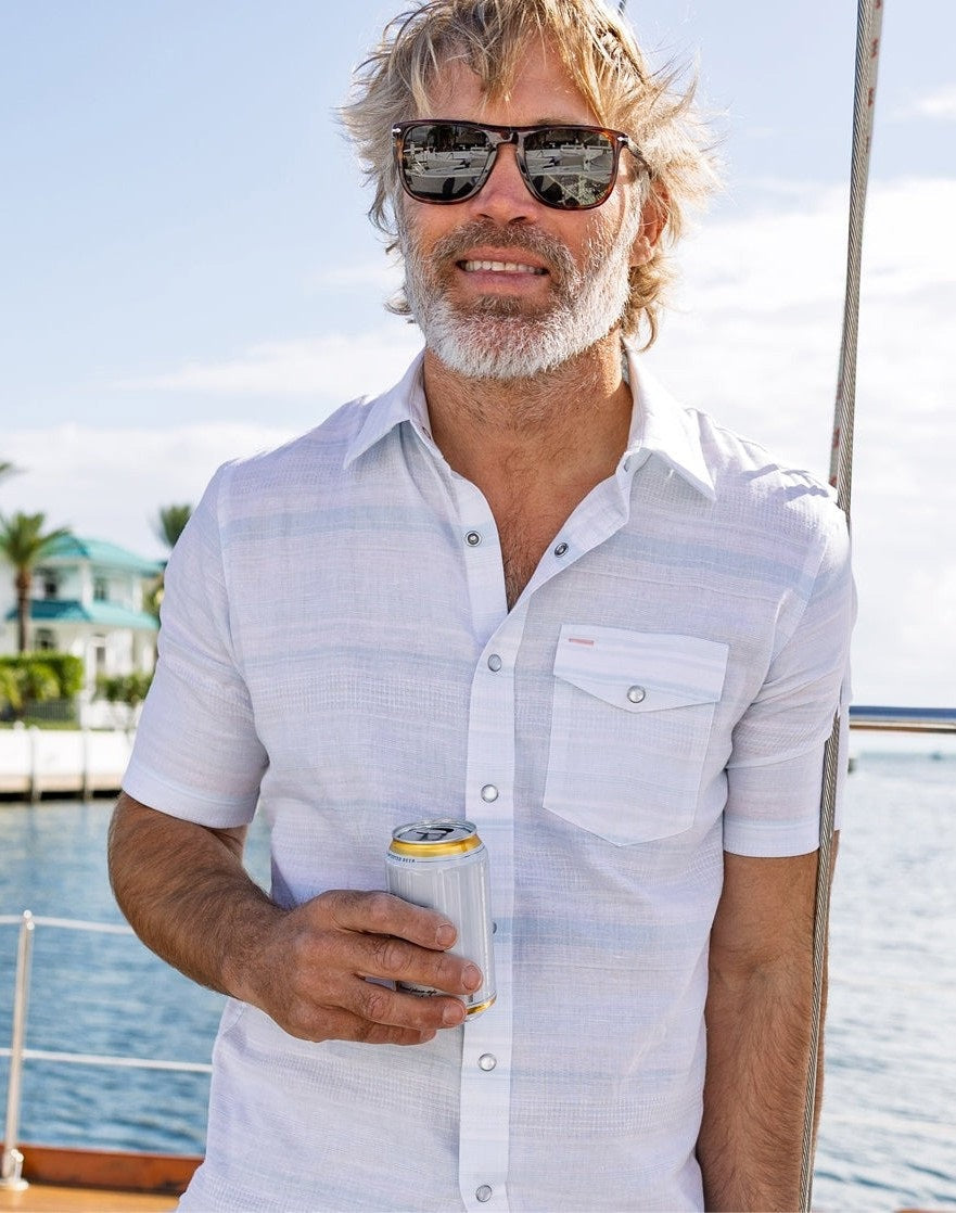 A man stands outdoors in sunlight wearing the CRIQUET HEMP STRIPE PEARL SNAP SHIRT IN LIGHT BLUE, featuring subtle horizontal stripes, short sleeves, a front pocket, and partial view of his bearded face. Brand: CRIQUET.