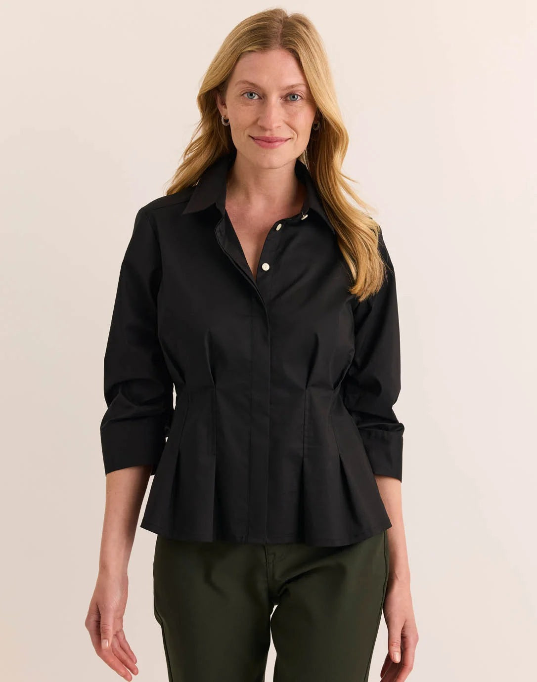 A woman with blonde hair is smiling at the camera while standing against a plain light background, wearing the HINSON WU - MARTINA 3/4 SLEEVE STRETCH POLISHED COTTON SHIRT IN BLACK paired with olive green pants.