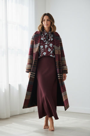 A woman stands indoors on light wooden flooring, barefoot and bathed in natural window light, effortlessly dressed for cold weather in the MIRTO 1956 - LONG PLAID WOOL COAT layered over a floral bow-tie blouse and a long burgundy skirt.