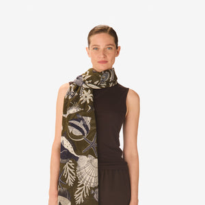 A person wears a white long-sleeve button-up shirt, an ocean-themed green, blue, and white sarong tied at the waist, and the INOUI - NEPTUNE SCARF IN KHAKI by INOUI, standing against a plain background.