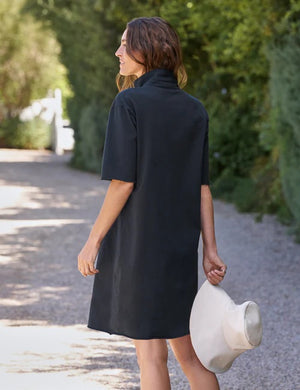 A woman in a loose, short-sleeve FRANK & EILEEN MINI EMMA Popover Henley Dress in Vintage Jersey British Royal Navy from the California Heritage Collection stands on a gravel path, smiling and looking down while holding a white wide-brimmed hat. Lush green foliage lines the background.