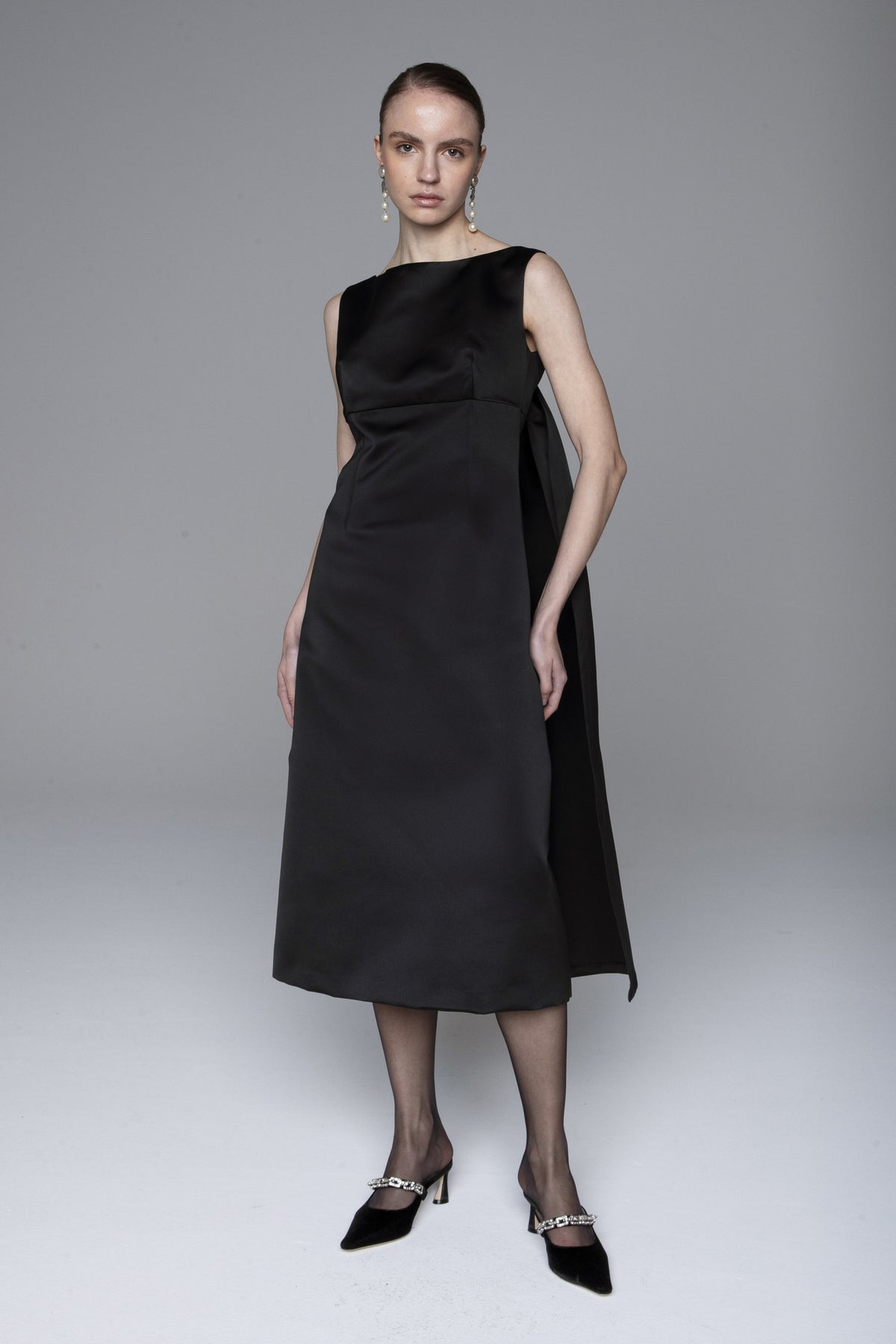 A woman stands facing away in the LAVI - NARA DRESS, a sleeveless black midi dress by LAVI reaching mid-calf. Her hair is in a neat bun, and she pairs it with black heels and drop earrings against a plain grey background.