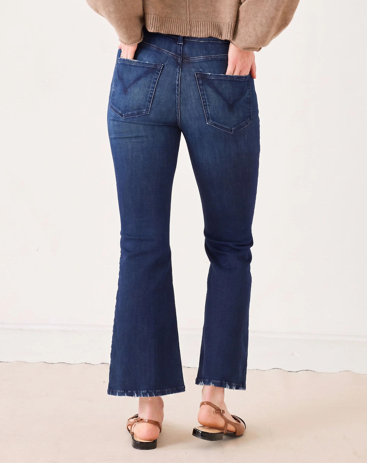 A person stands with hands in pockets against a plain light background, wearing MERSEA’s Murphy Mini Boot Jeans in Patriot—a pair of mid-rise, blue flare jeans crafted from four-way stretch denim—paired with a tan long-sleeve top, black pointed-toe flats, and gold bangles.