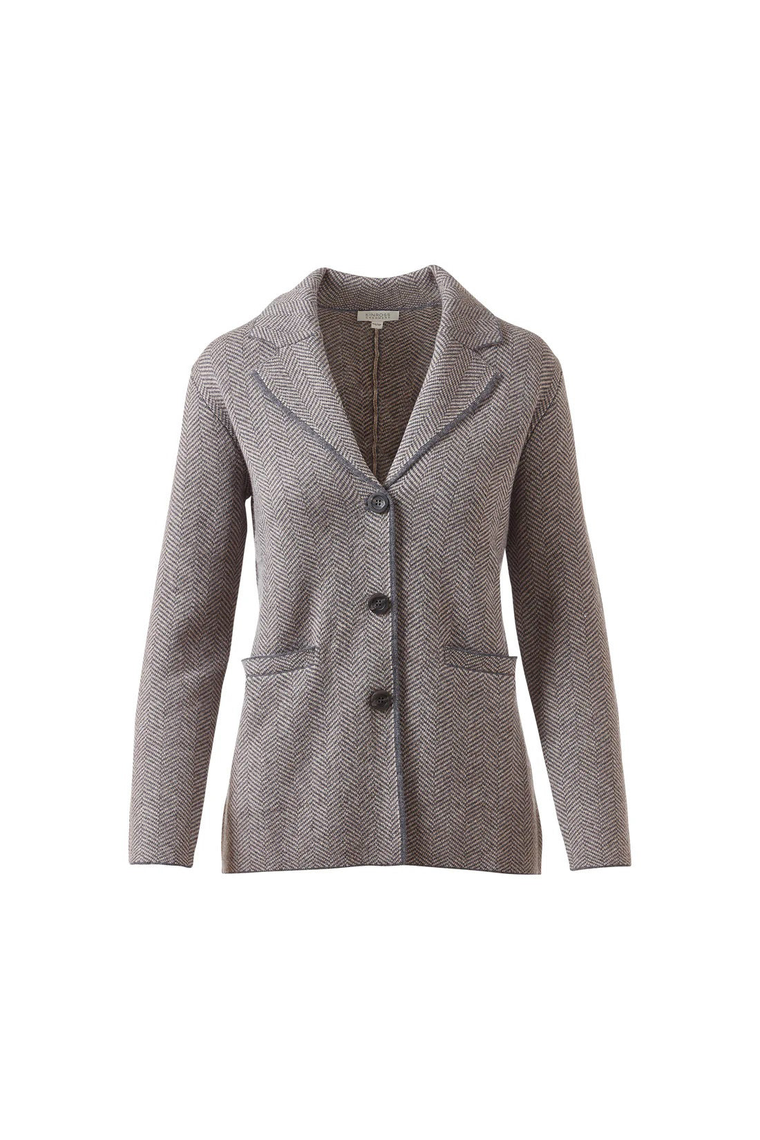 A woman with wavy brown hair wears the KINROSS CASHMERE Herringbone Notch Collar Knit Cardigan under a gray blazer with matching pants, standing against a white background with one hand in her pocket.