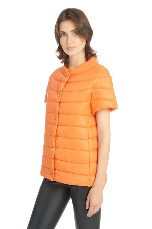 A person stands against a white background, showcasing the AUDREY PUFFER from PATTY KIM in vibrant orange. This short-sleeved jacket is ideal for the transition from fall to winter, paired stylishly with sleek black pants.