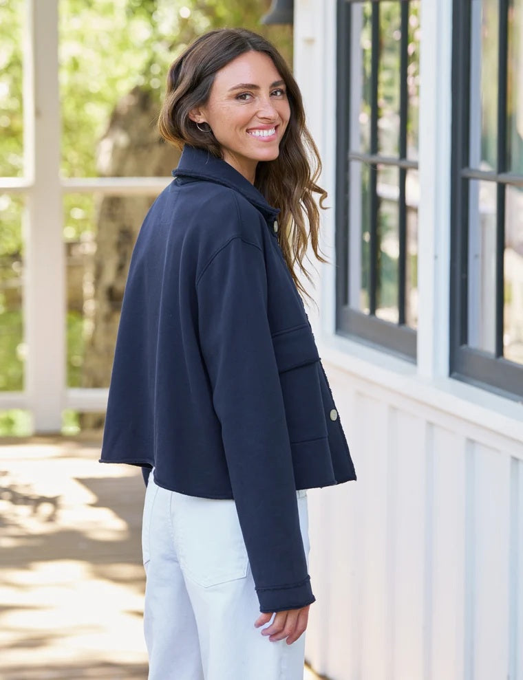 A woman with curly hair, wearing the FRANK & EILEEN Rowan Cropped Sweatshirt Utility Jacket in Triple Terry Classic Navy over a white top and light pants, leans against a wooden post and looks to the side.