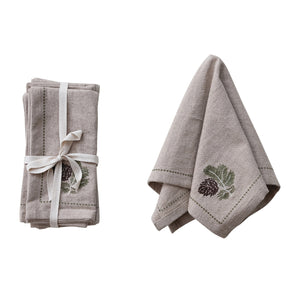 Four beige cotton NAPKIN WITH METALLIC THREAD EMBROIDERED BORDER by CREATIVE COOP, featuring embroidered pinecones and green leaves, are shown beside a pinecone branch on a white background.