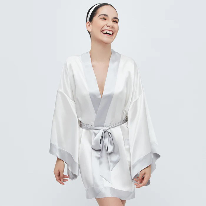The BRANCHE GEISHA Robe Silk in pink satin, adorned with white trim, hangs gracefully on a matching silk hanger. This elegant robe by BRANCHE features long, wide sleeves and is accentuated with a white sash tied in a bow around the waist, complete with mitered corners for an exquisite finish.