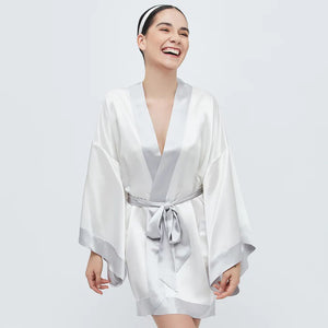 The BRANCHE GEISHA Robe Silk in pink satin, adorned with white trim, hangs gracefully on a matching silk hanger. This elegant robe by BRANCHE features long, wide sleeves and is accentuated with a white sash tied in a bow around the waist, complete with mitered corners for an exquisite finish.