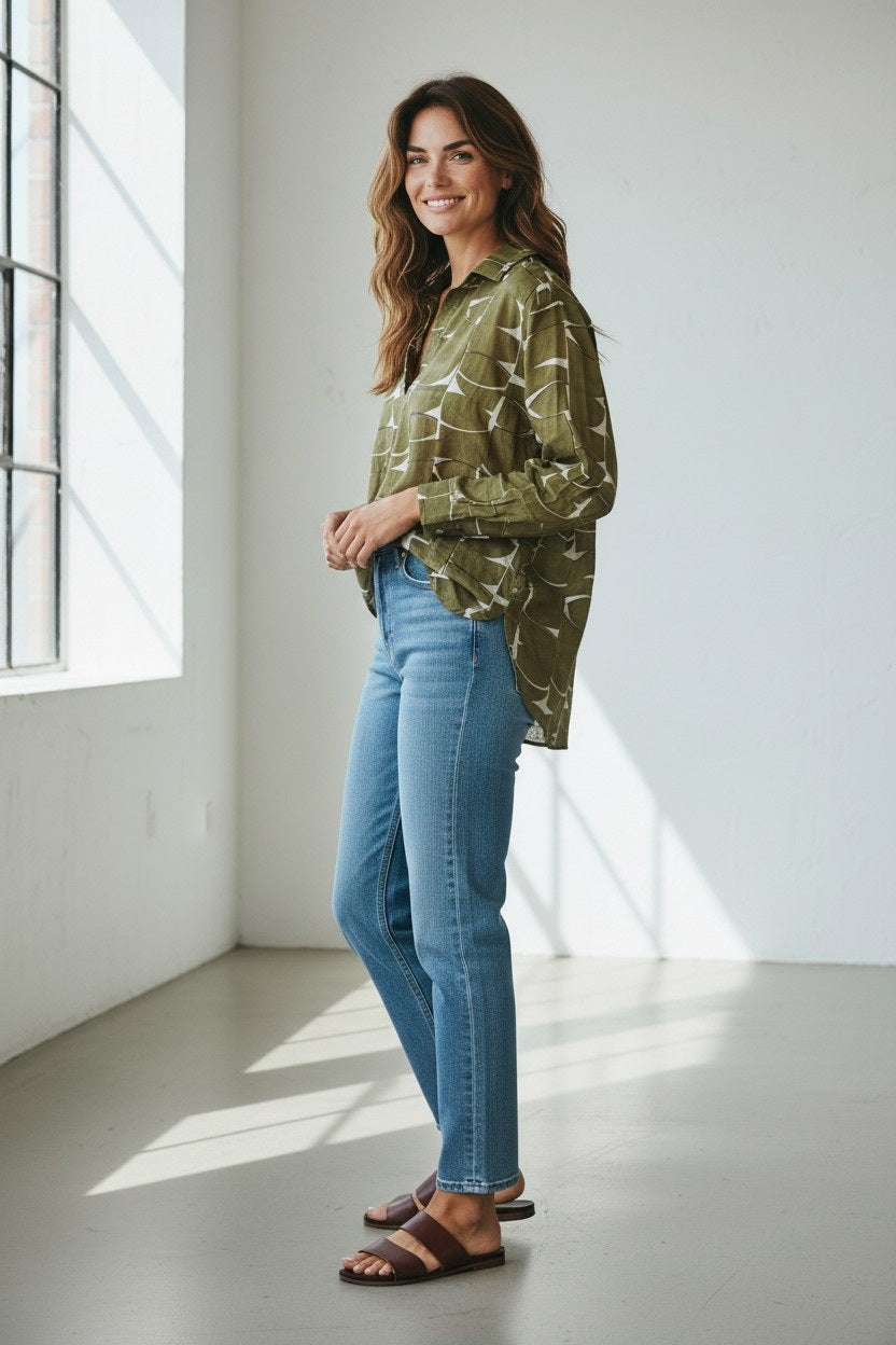 A woman stands in a sunlit room wearing the HARTFORD - CHORUS POPOVER SHIRT IN KHAKI by HARTFORD, paired with blue jeans and brown loafers, smiling with her hands in her pockets.