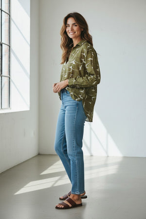 A woman stands in a sunlit room wearing the HARTFORD - CHORUS POPOVER SHIRT IN KHAKI by HARTFORD, paired with blue jeans and brown loafers, smiling with her hands in her pockets.