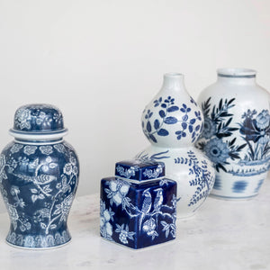 Four blue and white HAND PAINTED CERAMIC GINGER JAR vases by CREATIVE COOP, featuring floral and botanical motifs, are displayed on a white marble surface. The vases range in shape from classic jars to modern gourd styles.