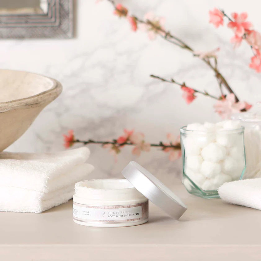 A clear plastic jar with a silver lid containing European Soaps, LLC Body Butter - Coconut, enriched with shea butter. The label displays &quot;Coconut&quot; and &quot;Body Butter | Beurre Corps,&quot; along with an illustration of coconut trees. The jar holds 6.7 fl oz (200 mL) of body butter to moisturize your skin.