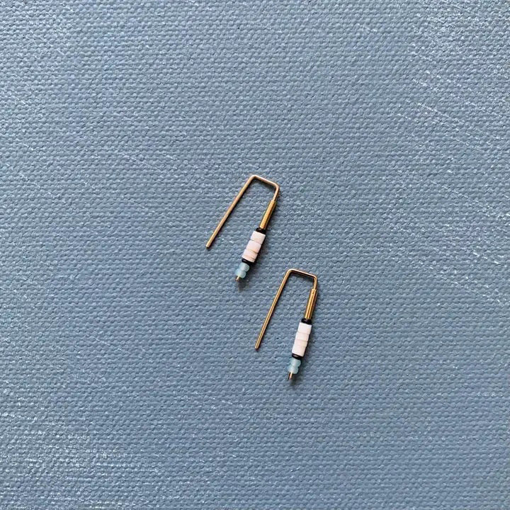 The ALICE RISE - LITTLE QUILL EARRINGS IN SHELL feature 14k gold filled rectangular bends threaded with pastel blue and white beads, displayed on a textured light blue background—an elegant handmade touch by ALICE RISE.