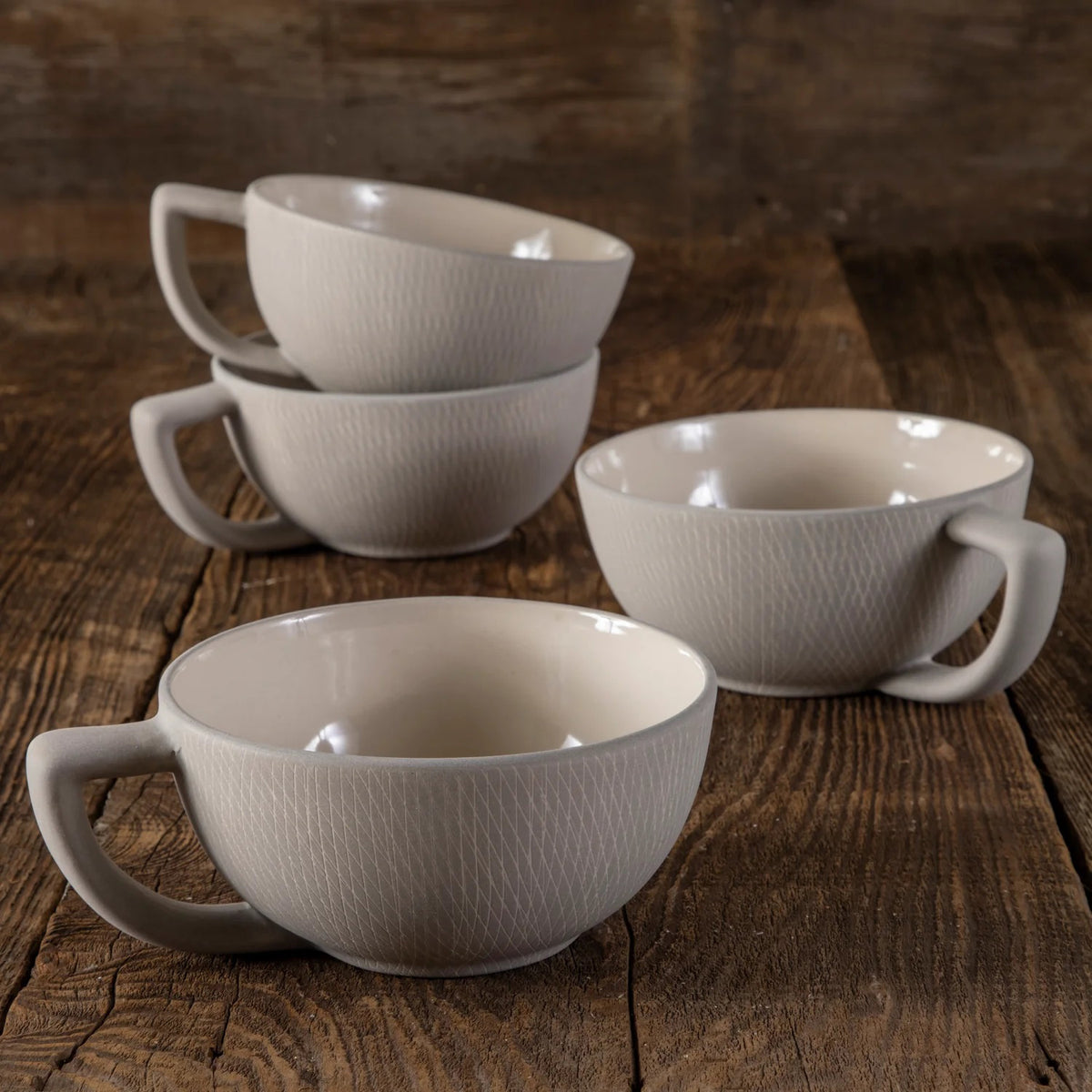 Four BE HOME - CROSSHATCH MUGS by BE HOME, INC, featuring textured light gray stone and handcrafted design, are stacked in pairs on a rustic wooden surface; two mugs appear in the foreground and two in the background.