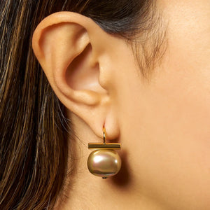 The CC & CO BY CATHERINE CANINO FOREVER FAVE PEBBLE PEARL EARRING IN BISCUIT features a pair of 14 karat gold earrings with round, iridescent pearls suspended beneath a sleek horizontal gold bar, displayed on a white background—a modern interpretation of classic pebble pearl earrings that effortlessly enhances any neutral beige jewelry collection.