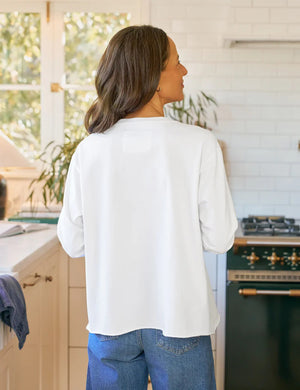 A woman with long brown hair wears the FRANK & EILEEN Quinn Long Sleeve Crewneck Tee in Heritage Jersey White, paired with blue jeans, standing in a sunlit kitchen with white tiles and a green stove, smiling softly.