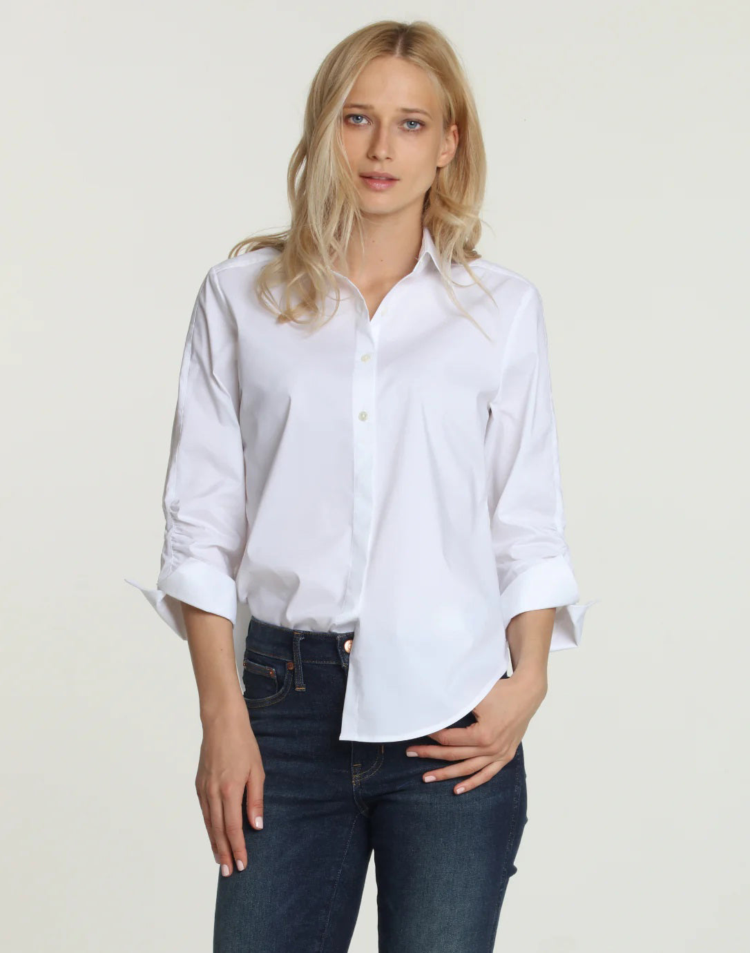 A person wearing the HINSON WU - ZOEY SHIRT, a finely tailored classic white piece with rolled-up sleeves, and paired with blue jeans, stands against a plain background with one hand in their pocket.