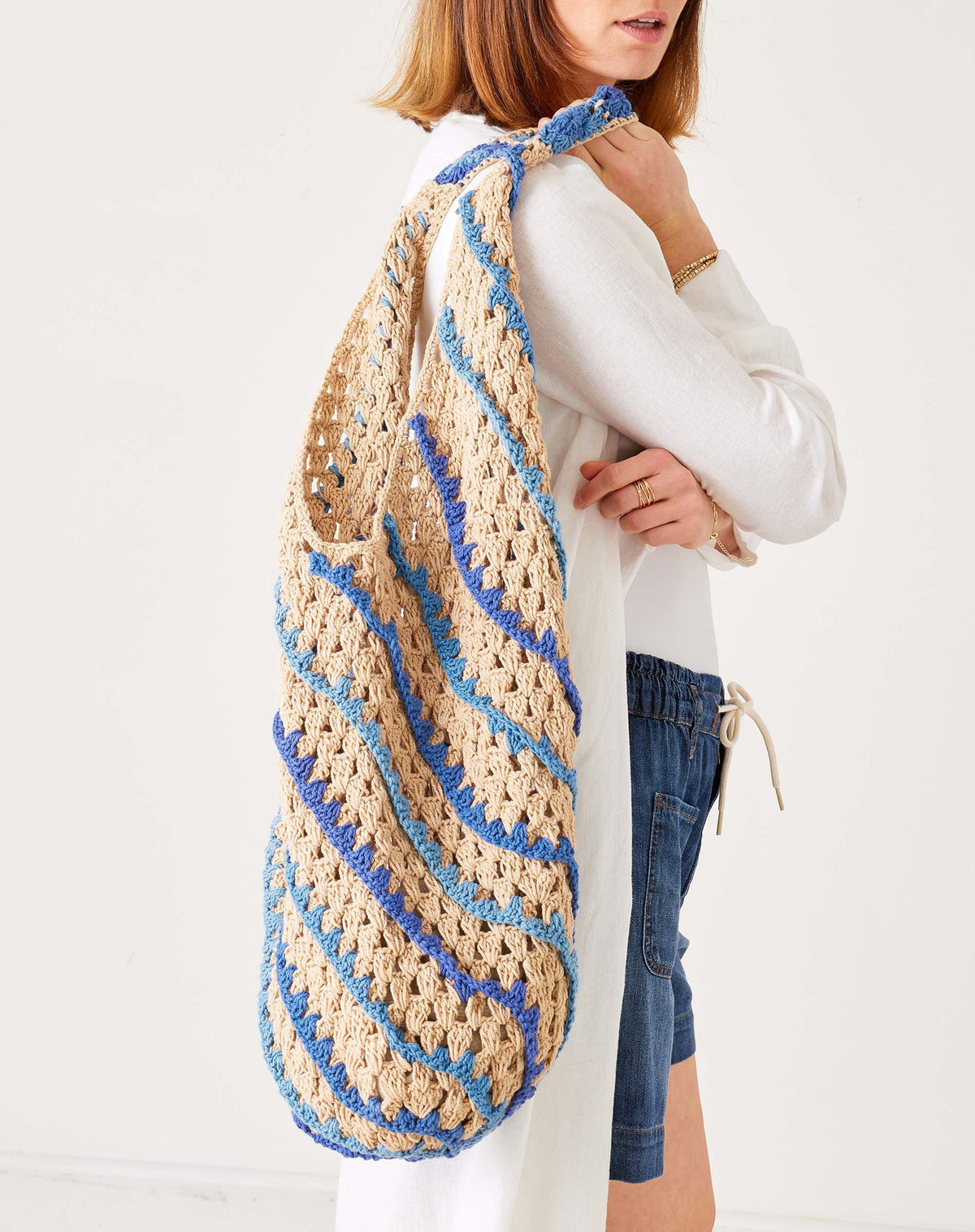 A person in white holds the MERSEA - CARLY CROCHET BOHO BAG by MERSEA, featuring large, colorful diagonal stripes of yellow, pink, red, green, and purple against a plain light background.