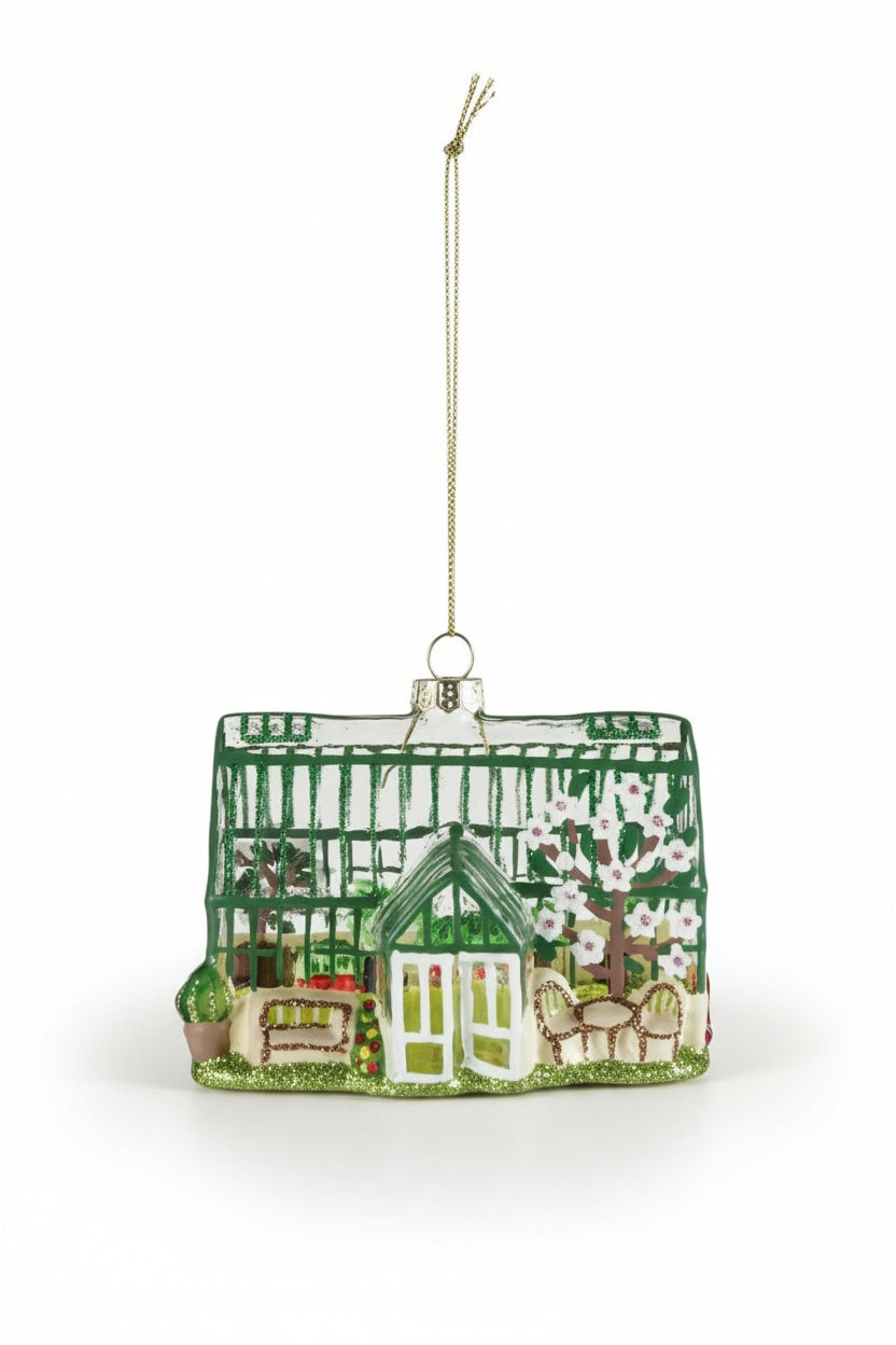 Hand-painted GREENHOUSE ORNAMENT WITH GLITTER featuring interior plants, furniture, and white flowers, hanging from a gold string.