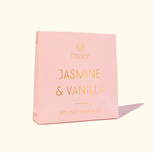 A pink package labeled “MUSEE - JASMINE & VANILLA MINI BATH SALT SOAK” by MUSEE, with a small pile of white bath salts spilled next to it, highlights the soothing benefits of natural bath products. Relax and unwind as you enjoy the calming jasmine vanilla aroma in your Epsom salt bath for ultimate tranquility.