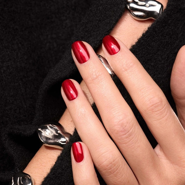 A hand with shiny, iridescent red nails holds a matching bottle labeled “MANUCURIST - GREEN™ NAIL POLISH IN RED HIBISCUS” by MANUCURIST INC against brown skin. This luxurious vegan, cruelty-free nail polish delivers vibrant color and a flawless finish.