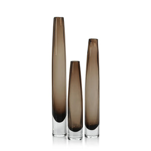 The ZODAX TATE Slim Glass Taupe Vase - Tall features a narrow cylindrical design in smoky brown glass with a clear base, ideal for display on a glass shelf against a plain white background.