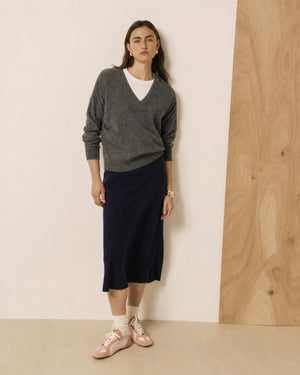 A woman with long brown hair and a neutral expression stands against a two-tone wall, wearing a grey sweater layered over a white shirt, white socks, pink shoes, and the HARTFORD - JIMA Bias Cut Skirt in Night by HARTFORD, effortlessly showcasing chic style with this navy midi skirt.