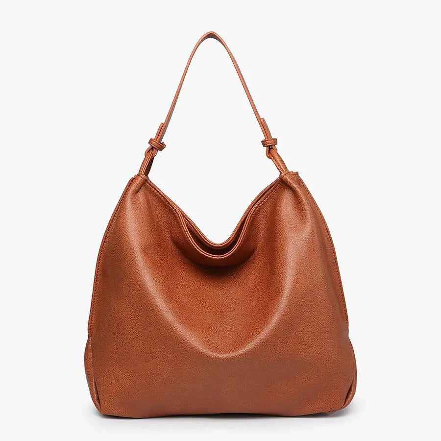 The JEN &amp; CO RUSTIC TOTE is crafted from brown vegan leather and features a relaxed, slouchy shape. Its simple and elegant design includes a single strap attached at both sides, along with an inner compartment for added functionality, all set against a plain light background.