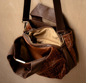 The DANIELLA LEHAVI EVELINE Satchel in Brown Mosaic, featuring double handles, a croco-inspired texture, and an adjustable shoulder strap, hangs on a wall hook against a light wooden background.