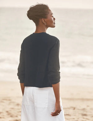 A woman stands on the beach, looking down and smiling as she wears the FRANK & EILEEN Jackie T-Shirt Cardigan in Vintage Jersey British Royal Navy, gently touching her collarbone with her left hand. The background is softly blurred with waves in view.